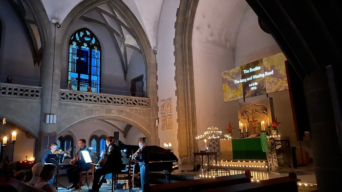 In der Kirche spielen zwei Musiker auf einem Klavier, umgeben von Kerzenlicht und hohen Fenstern.