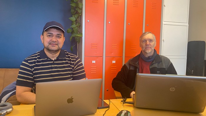 Zwei Männer sitzen an einem Tisch mit Laptops, einer trägt ein gestreiftes Hemd und eine Mütze, der andere eine Jacke.