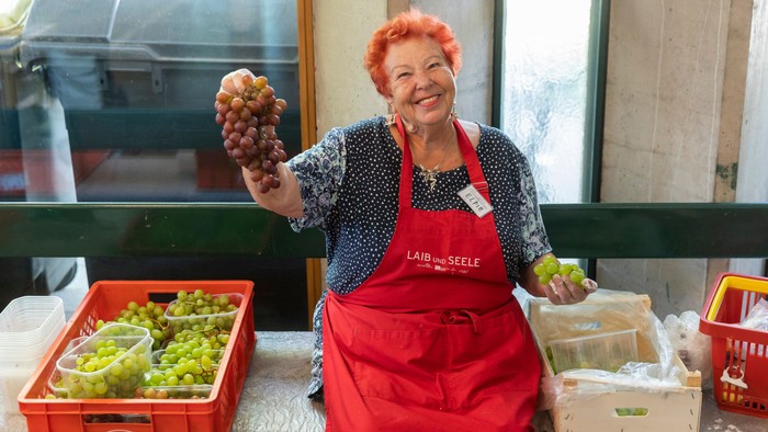 Eine ältere Frau in einer roten Schürze hält Trauben in der Hand und steht vor Kisten mit Obst.