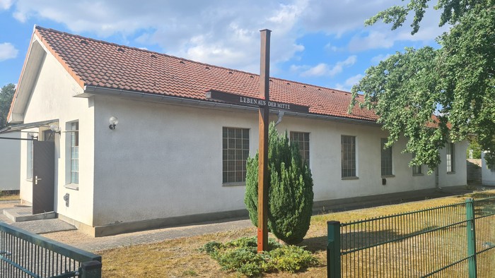 Kleines weißes Gebäude mit rotem Dach und Schornstein, umgeben von einem Zaun und Grünfläche