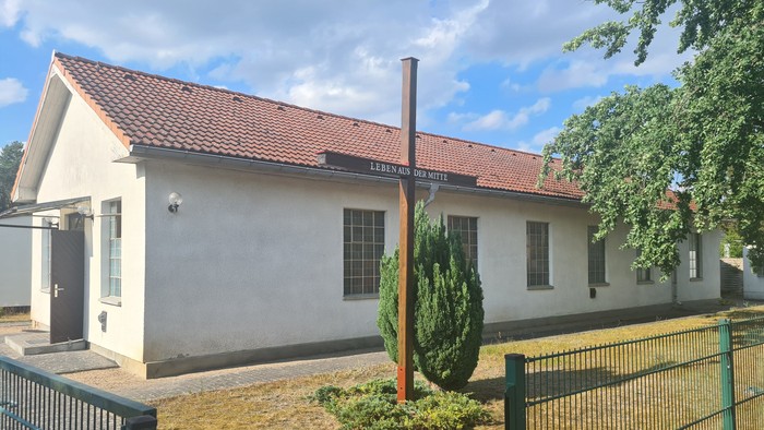 Kleines weißes Gebäude mit rotem Dach und Schornstein, umgeben von einem Zaun und Grünfläche