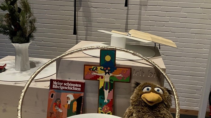 Auf einem Tisch stehen eine Schale, eine Puppe, ein Buch und ein Kreuz vor einer weißen Ziegelwand