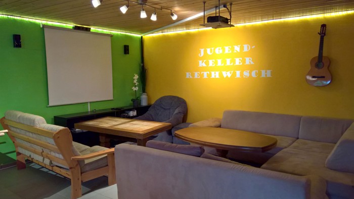 Gemütliches Wohnzimmer mit beigen und grauen Möbeln, Gitarre an der Wand, Projektor und leuchtender Deckenbeleuchtung