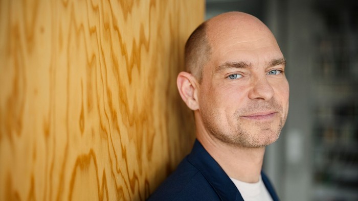 Mann mit Glatze lehnt an Holzpaneel