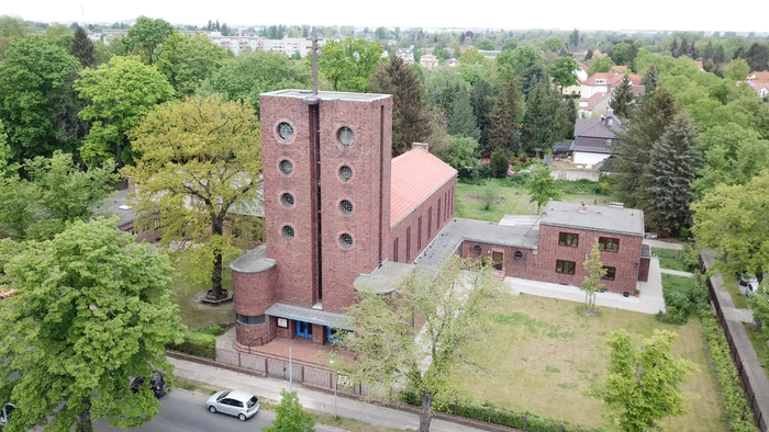 Das Bild zeigt ein großes Ziegelgebäude mit markantem Turm inmitten von Bäumen und Wohnhäusern.