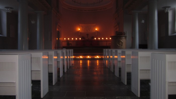 En mørk kirkeinterior med lysende lys i baggrunden og hvide bænkefløje på hver side.