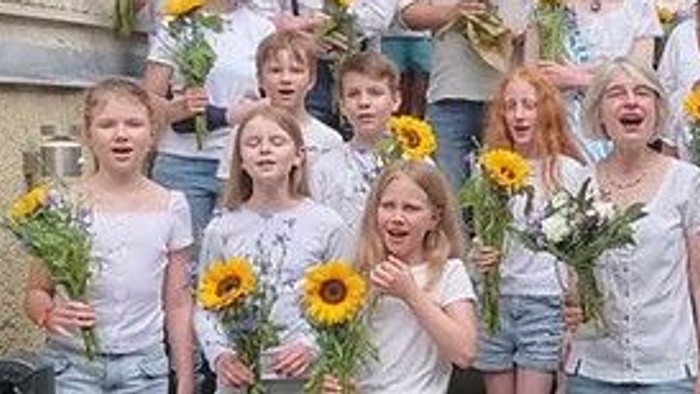 Eine Gruppe von Menschen, darunter Kinder und Erwachsene, posiert mit Sonnenblumen vor einem Gebäude.