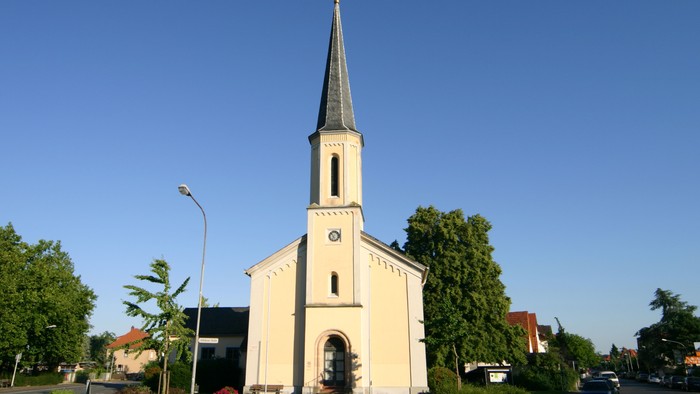 Eine malerische Kirche mit hohem Kirchturm vor einem klaren blauen Himmel, umgeben von Grünflächen und Wohngebäuden.