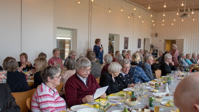 En gruppe mennesker sidder ved bord i en lys og rummelig lokalitet, muligvis ved en fest eller sammenkomst.