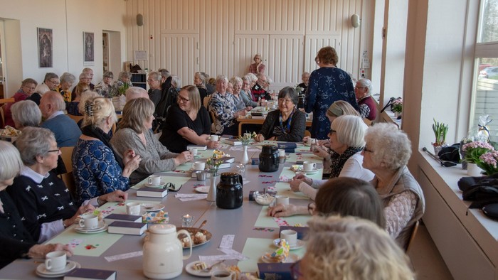 En gruppe af ældre folk sidder ved lange bord og spiser. Der er servisebåde og mad på bordet.