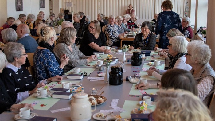 En gruppe pensionister sidder ved lange bord og spiser te og kage.