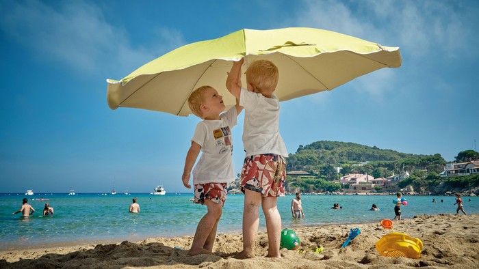 Zwei Kinder stehen am Strand unter einem gelben Regenschirm.