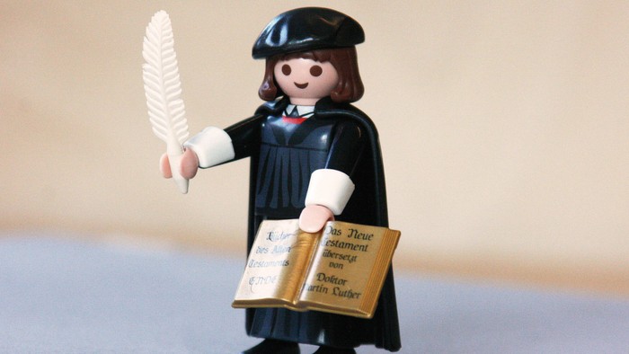 Eine kleine Figur, gekleidet als Gelehrter, hält eine Feder und ein aufgeschlagenes Buch.