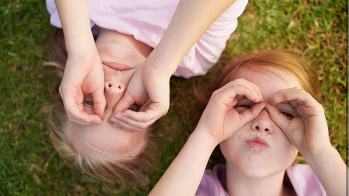Zwei Kinder bedecken sich spielerisch gegenseitig die Augen, während sie auf dem Gras liegen und lustige Grimassen schneiden.