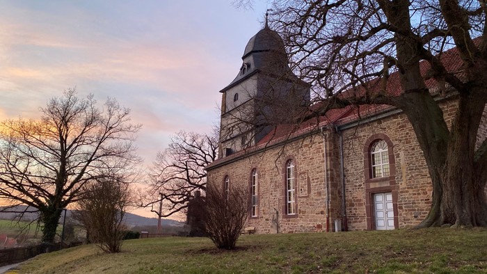 Steinerne Kirche mit Turm, umgeben von kahlen Bäumen und einer grasbewachsenen Fläche, unter einem farbenfrohen Himmel.