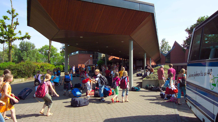 Kinder mit Fahrrädern und Rollern stehen vor einem Bus an einer Bushaltestelle.