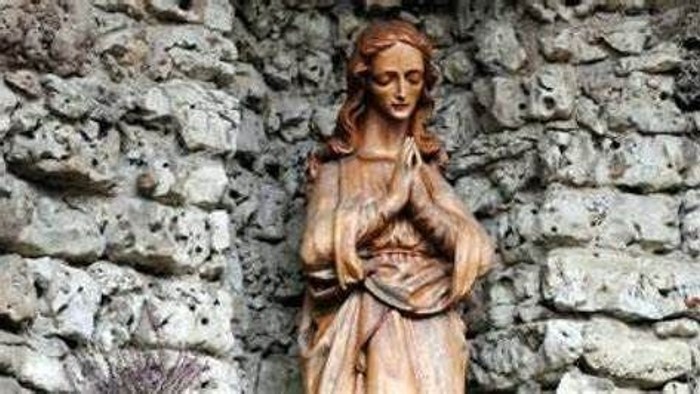 Die Statue einer Frau mit gefalteten Händen vor einer steinernen Wand.