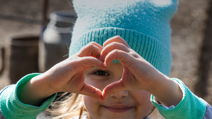 Et smilende barn med en blå hat laver et hjerte med hænderne og beder om hjælp til ukrainske børn.