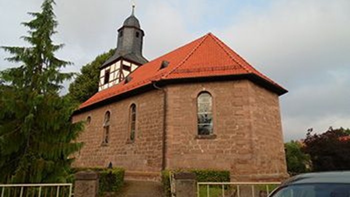 Kirchengebäude mit rotem Dach und Turm, umgeben von Bäumen und einem Auto davor