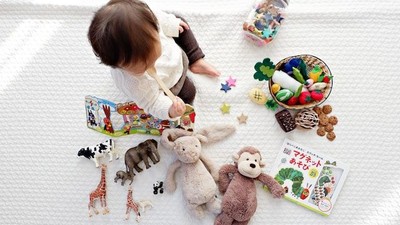 Ein Baby sitzt auf einer Decke umgeben von Spielzeugen wie Tieren, Büchern und einem Behälter mit kleinen Gegenständen.