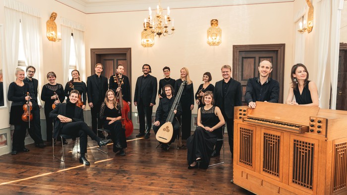 Ein Orchester posiert in einem eleganten Raum mit Kronleuchtern und Türen.