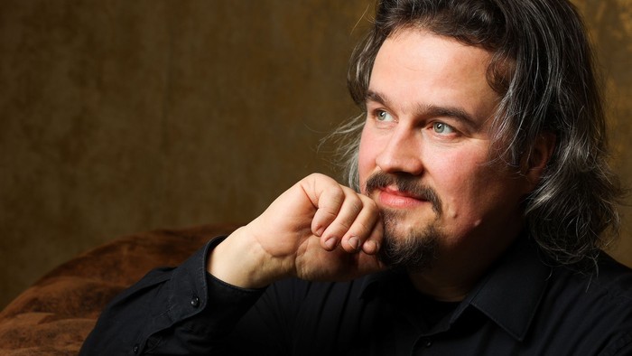 Ein Mann mit langem Haar und Bart sitzt nachdenklich auf einem braunen Sofa.