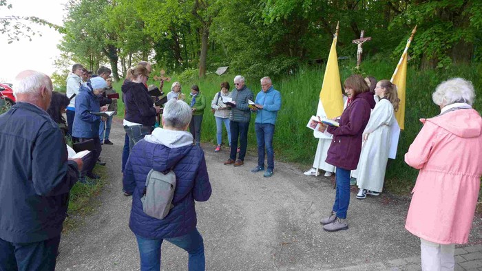 Eine Gruppe von Menschen steht im Freien und hält Bücher, einige tragen Regenmäntel und Schirme