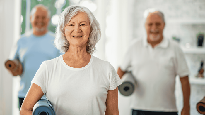 Eine ältere Frau lächelt in einem Fitnessstudio, umgeben von anderen Senioren mit Yogamatten.