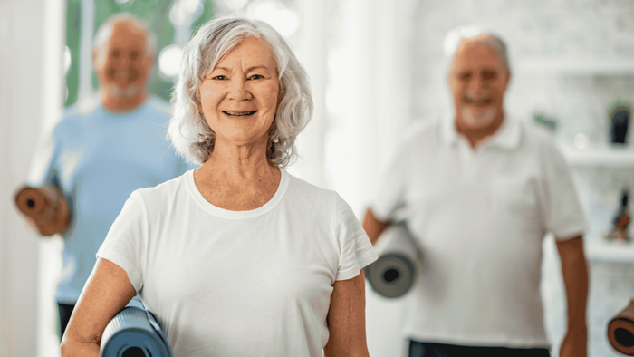 Eine ältere Frau lächelt in einem Fitnessstudio, umgeben von anderen Senioren mit Yogamatten.