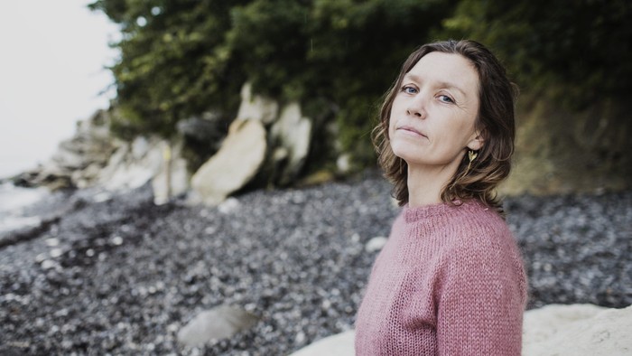 En person i en lyserød sweater står eftertænksomt på en stenet strand nær træer.
