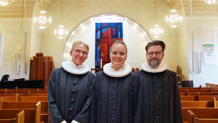 Tre gejstlige i kjortler poser sammen inde i en kirke med træbænke og farvede glasvinduer.