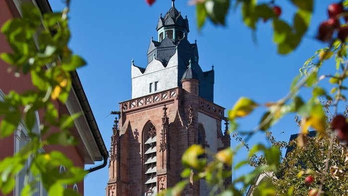 Turm einer Kirche mit Uhr und spitzen Dach, umgeben von Bäumen und rotem Gebäude.
