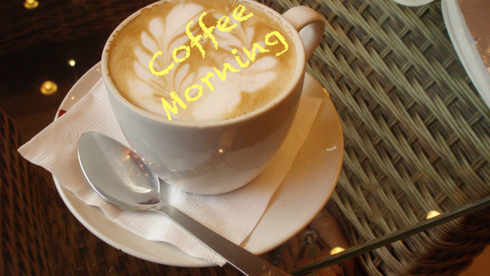 Cup of coffee with "Coffee Morning" written in foam, spoon on saucer