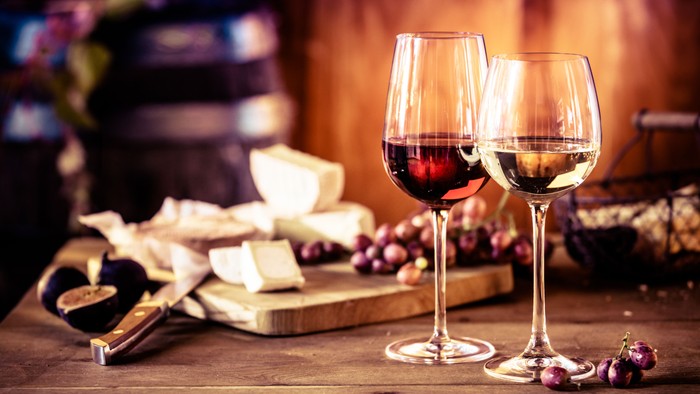 Im Vordergrund ein Glas Weißwein und ein Glas Rotwein. Im Hintergrund eine Käseplatte mit Trauben.