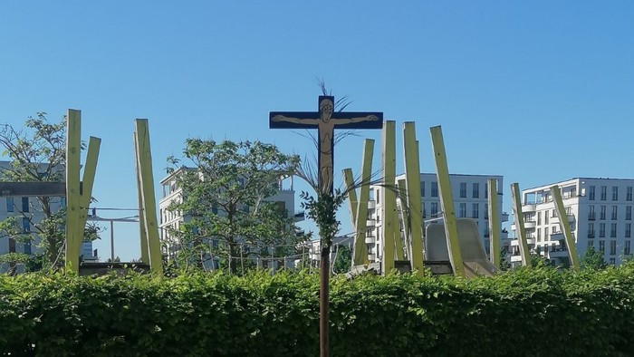 Ein hölzernes Kreuz steht auf einem Betonpfahl im Freien, umgeben von Büschen und Gebäuden im Hintergrund.