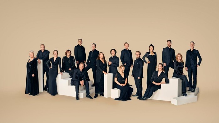 En gruppe af 14 personer i mørke klæder poser på en beige baggrund.