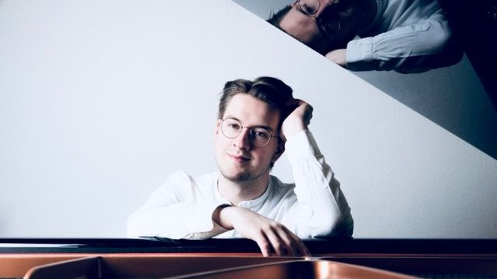 Ein Mann mit Brille lehnt an einem weißen Objekt und schaut zur Seite.