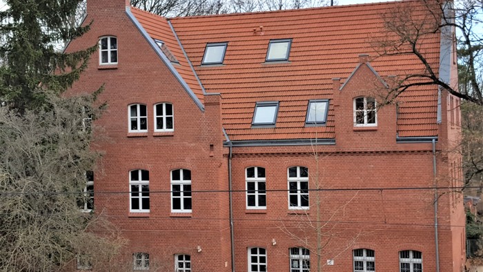 Das Bild zeigt ein zweistöckiges rotes Backsteingebäude mit mehreren Fenstern und einem roten Ziegeldach.