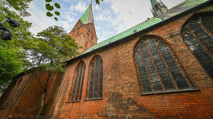 Historische Backsteinkirche mit hohen Türmen und bogenförmigen Buntglasfenstern, die sich vor einem grünen Dach abheben.