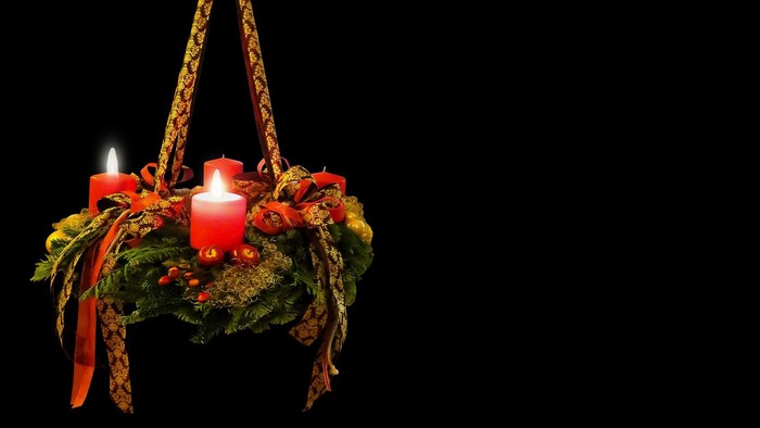 Ein Adventskranz mit vier brennenden Kerzen, geschmückt mit Tannenzweigen und bunten Bändern, hängt an einem schwarzen Hintergrund.
