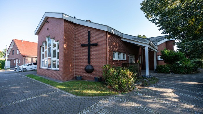 Ziegelkirche mit Kreuzsymbol an der Fassade, umgeben von Bäumen und anderen Gebäuden.
