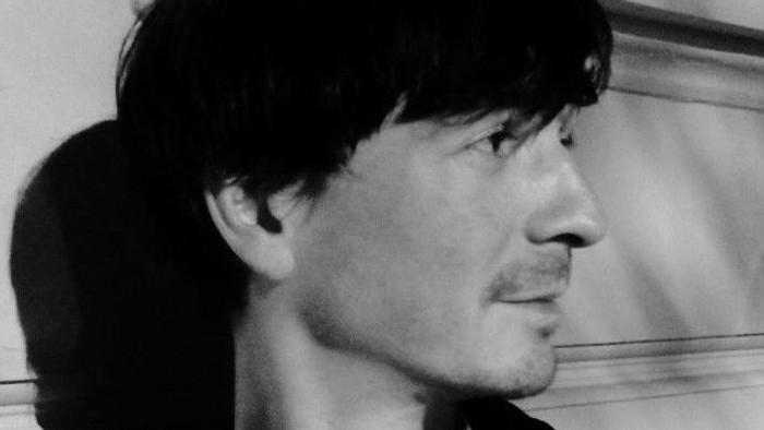 Schwarzes und weißes Streifenhemd, dunkle Jacke, ernstes Gesicht.