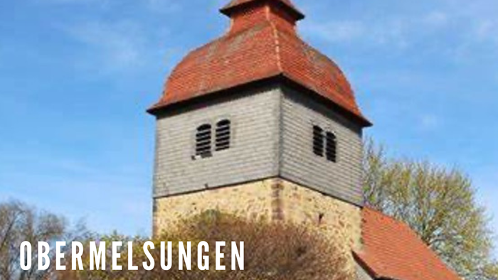 Kirchturm mit rotem Dach und kleinen Fenstern, umgeben von Bäumen unter blauem Himmel.