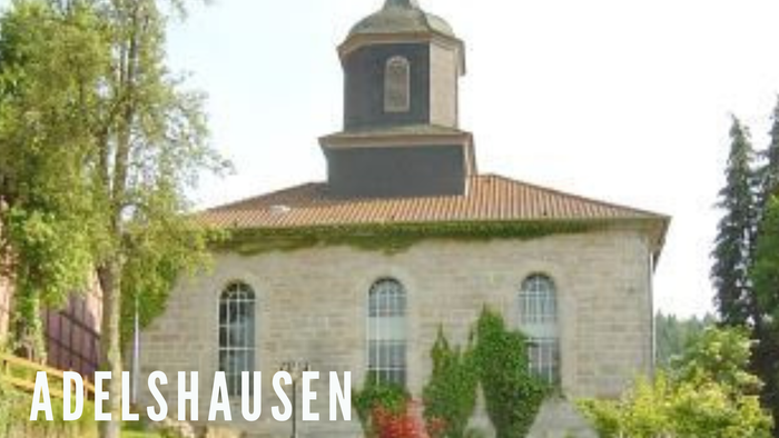 Steinkirche mit Glockenturm und Rundbogenfenstern, umgeben von Grünflächen.