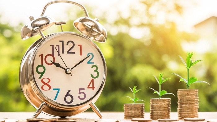 Ein Wecker mit bunten Zahlen steht neben drei Topfpflanzen auf einem Holzbalkon.