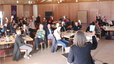 Ein großer Raum mit vielen Menschen an Tischen, einige sitzen, andere stehen und reden.