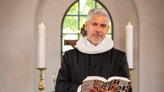 Mand i sort kåbe, der holder en bog i en kirke.