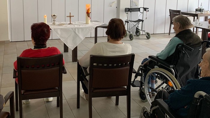 Die Gruppe versammelt sich in einem ruhigen Raum mit einem kleinen Altar, einige sitzen auf Stühlen, eine Person im Rollstuhl.