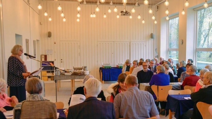 En gruppe mennesker sidder i en lys og rummelig sal og lytter til en tale.