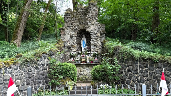 Ein steinerner Schrein mit Marienstatue in einem grünen Garten mit Bänken und Blumen.